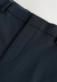 Gros plan de la ceinture d'un pantalon bleu marine foncé avec passants de ceinture et une poche sur la couture latérale.