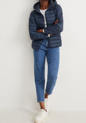 Person stående iført en navy dunjakke, hvid skjorte, blå jeans og hvide sneakers mod en ensartet lys baggrund.