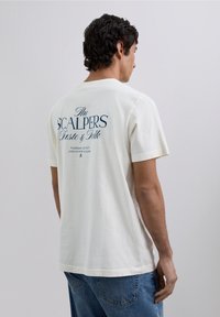 Cremefarbenes Baumwoll-T-Shirt mit kurzen Ärmeln. Auf der Rückseite befindet sich dunkelblauer Textdruck mit Markennamen und Veranstaltungsdetails.