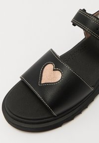 Sorte lærsandaler med en bred rem, med et metallisk rosa hjerte-design, forsterkede sømmer og en teksturert såle.