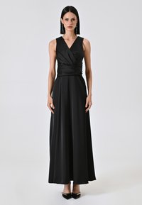 Schwarzes ärmelloses Kleid aus glattem Stoff, mit einem wickelnden Oberteil-Design und einem langen, fließenden Rock. Kombiniert mit spitzen schwarzen Absätzen.
