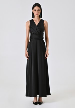 Schwarzes ärmelloses Kleid aus glattem Stoff, mit einem wickelnden Oberteil-Design und einem langen, fließenden Rock. Kombiniert mit spitzen schwarzen Absätzen.