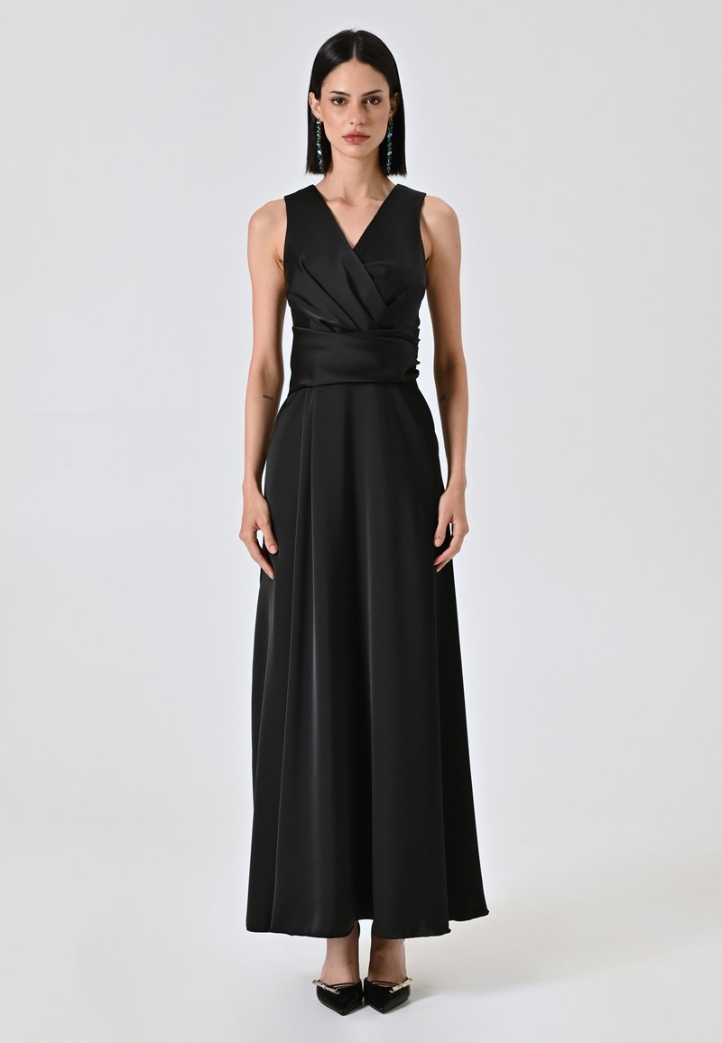 Robe noire sans manches en tissu lisse, présentant un design de corsage drapé et une longue jupe fluide. Associée à des talons aiguilles noirs.