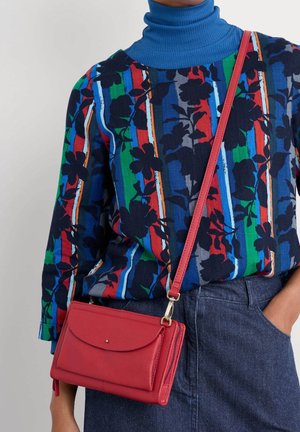Person trägt bunten gestreiften floralen Pullover mit blauem Rollkragen, dunkelblaue Jeans und trägt eine kleine rote Umhängetasche.