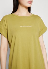 Olivgrünes T-Shirt aus weicher Baumwolle, mit aufgerollten kurzen Ärmeln und weißer Aufschrift "MSHCPOPENHAGEN" auf der Brust.