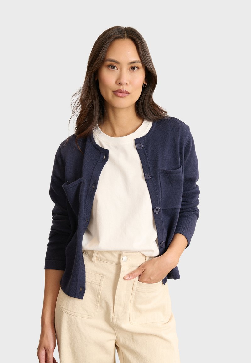 Mörkblå cardigan med långa ärmar, knäppning fram och en liten bröstficka, stylad över en vit t-shirt och krämfärgade byxor.