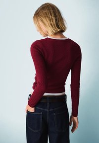 Bordeaux kabelgebreide trui met lange mouwen en een aangesloten ontwerp, gecombineerd met donkere spijkerbroek en een zwarte riem. Achteraanzicht.