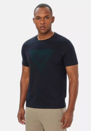 Uomo che indossa una t-shirt nera con il logo Guess e pantaloni beige in piedi davanti a uno sfondo bianco uniforme.