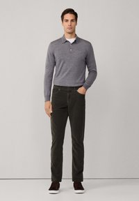 Man staand tegen een effen achtergrond, gekleed in een grijze lange mouwen polo, donkere groene broek en donkere sneakers met witte zolen.