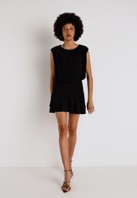 Vestido preto com um corpete solto e drapeado, mangas curtas e saia com babados. Modelo está a usar sapatos de salto verdes-azeitona.