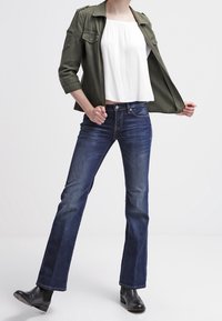 Giacca militare verde oliva, top bianco scollato sulle spalle, jeans a zampa d'elefante blu scuro e stivaletti neri alla caviglia. La modella posa con una mano sulla giacca.