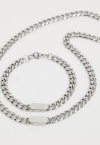 Sølv kæderekæde og armbånd sæt, der har sammenflettede led, en glat overflade og flade ovale låse.