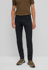 Musta puuvillapantsit slim-fit-mallissa, jossa on viisi taskua, vetoketjulukko ja hienovarainen logotieto. Yhdistetty ruskean T-paidan ja vaelluskengillä.