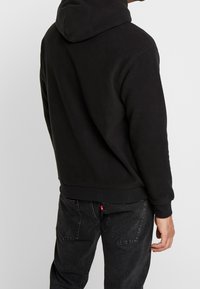 Felpa nera in pile con tasca a marsupio, cappuccio con cordino, e polsini e orlo a costine. Indossata con jeans neri caratterizzati da un dettaglio con etichetta rossa.