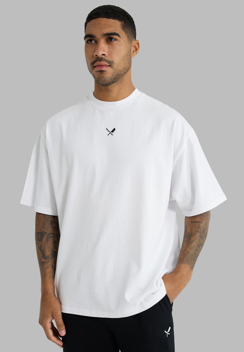 Homme tatoué portant un t-shirt blanc ample et un pantalon noir, tous deux présentant de petits logos croisés de couteau et fourchette sur un fond gris uni.