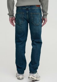 Jeans din denim de culoare albastru închis, cu o croială dreaptă, au două buzunare la spate, o etichetă din piele la talie și sunt asortați cu adidași deschiși la culoare.