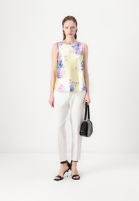 Blusa sin mangas amarilla con estampado floral y acentos morados, combinada con pantalones blancos ajustados y tacones negros con tiras, sosteniendo un pequeño bolso negro.