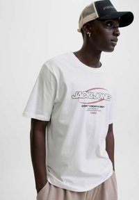 Jauns vīrietis, tērpies baltā Jack & Jones T-kreklā un melnā, bēšā cepurē, stāv ar rokām kabatās, vērsts pa labi.
