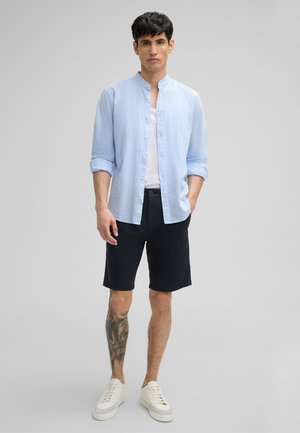 Light blue button-up shirt with a mandarin collar, paired with dark shorts and white sneakers. Short sleeves and a casual fit.