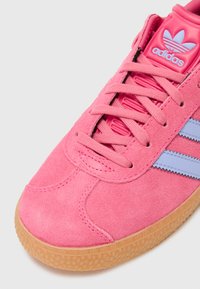 Zapatilla de ante rosa con rayas azul claro y cordones a juego. Presenta una suela de goma texturizada y marca en la lengüeta.
