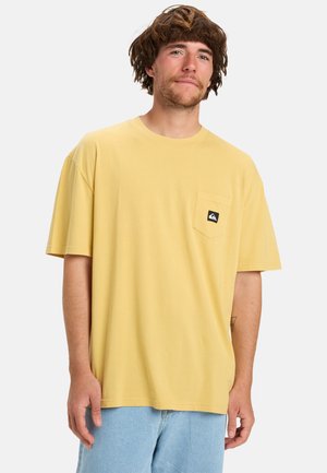 Gul bomulds T-shirt med korte ærmer, der har en lomme foran og et sort logo patch. Båret med lyseblå denim shorts.