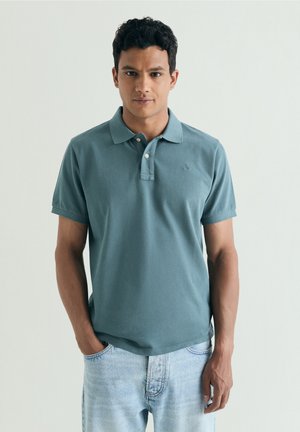 Ung mand stående med hænderne i lommerne iført en grønblå polo og lyseblå jeans mod en ensfarvet baggrund.