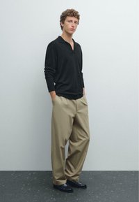 Svart långärmad polo med krage, kombinerad med beige plisserade byxor och svarta skor, stående mot en neutral vägg.