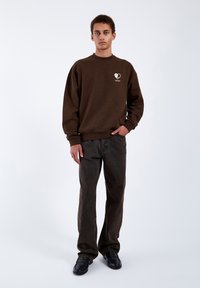 Felpa marrone con un logo a forma di cuore bianco, abbinata a jeans denim scuri e scarpe nere. Design semplice, vestibilità rilassata e polsini a costine.