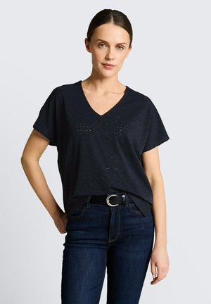 Femme aux cheveux foncés en chignon bas porte un haut noir à manches courtes avec col en V et un jean bleu foncé avec une ceinture noire sur un fond uni.