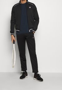 Czarna kurtka sportowa z białymi paskami, granatowa koszulka, czarne spodnie z detalem logo i czarne trampki, trzymająca rakietę tenisową.