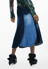 Jupe en denim composée de panneaux bleu foncé et bleu clair, coupe évasée et ourlet brut. Associée à des bottines noires brillantes.