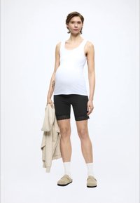 Λευκό αμάνικο τοπ, μαύρο σορτς με δαντελωτή επένδυση, μπεζ ζακέτα κρεμασμένη στο χέρι, ανοιχτόχρωμες κάλτσες και παπούτσια τύπου slip-on. Το μοντέλο στέκεται μπροστά σε απλό φόντο.