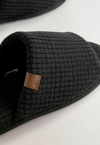 Des chaussons noirs avec un motif gaufré texturé, arborant une étiquette en cuir marron et un design courbé à bout ouvert.