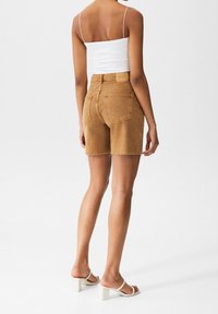 Shorts en denim brun avec un ourlet effiloché, deux poches arrière et des détails de couture visibles. Portés avec un top à bretelles blanc et des sandales claires.