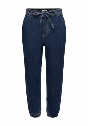 Kobieta ubrana w ciemnoniebieskie dżinsowe spodnie jogger z wysokim stanem i elastycznymi mankietami, żółty top z długim rękawem oraz czarne klapki.