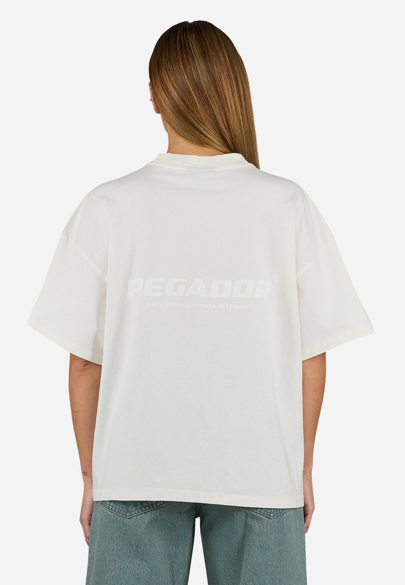 Bijela oversized majica kratkih rukava s odvažnim "PEGADOR" logotipom na leđima, izrađena od mekanog pamučnog materijala.