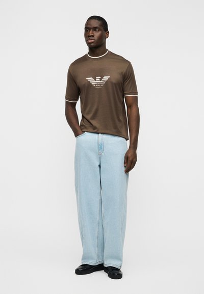 Καφέ κοντομάνικο μπλουζάκι με λευκό γιακά και λογότυπο, συνδυασμένο με ανοιχτό μπλε τζιν σε χαλαρή γραμμή και μαύρα παπούτσια.