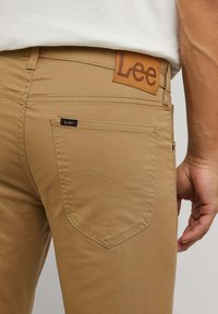 Bruine broek van een katoenmix, met een lederen label met het "Lee"-logo, een achterzak met een klein zwart labeltje en een gebogen stiksel detail.