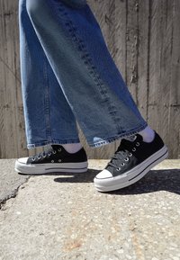 Schwarze Canvas-Sneaker mit weißen Gummisohlen und Zehenkappen, kombiniert mit weit geschnittenen blauen Jeans, vor einer strukturierten Betonoberfläche.