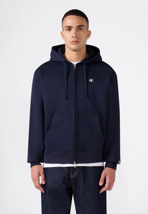 Junger Mann, der einen marineblauen Kapuzenpullover mit Reißverschluss und kleinem Logo auf der Brust sowie dunkle Jeans trägt, vor einem schlichten Hintergrund stehend.
