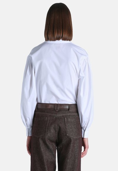 Femme aux cheveux bruns raides portant une chemise blanche à manches longues rentrée dans un pantalon marron foncé avec une ceinture en cuir, vue de dos.