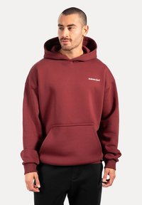 Bordowy hoodie z przednią kieszenią, kapturem ze sznurkiem i ściągaczami rękawów. Posiada małe białe logo "Smilodax Sport" na piersi.
