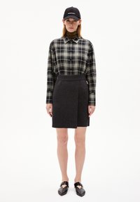 Camicia a maniche lunghe a quadri neri e verdi, gonna avvolgente grigia e strutturata, berretto nero con testo "PERMANENT" e scarpe flat nere.