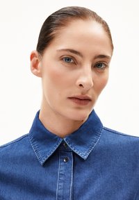 Camicia di denim blu profondo con colletto strutturato, caratterizzata da una chiusura a bottoni e una texture liscia, che mette in evidenza i dettagli delle cuciture.