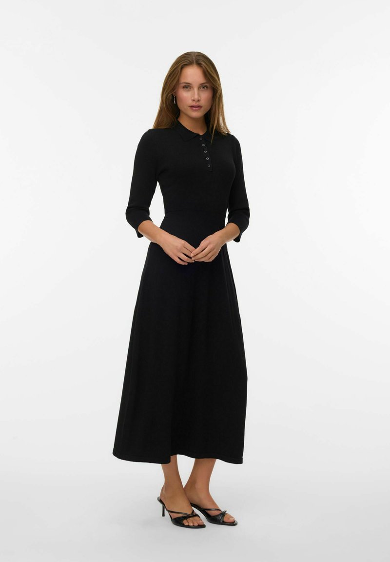 Vestito lungo nero a maniche lunghe con colletto e pattina di bottoni, corpino aderente, gonna svasata, texture liscia, abbinato a tacchi neri con cinturino.