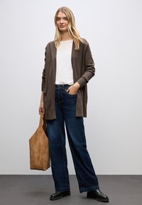 Brązowy kardigan z prążkowanymi mankietami na białej bluzce, połączony z szerokimi niebieskimi dżinsami i czarnymi botkami. Trzyma beżową zamszową torbę.