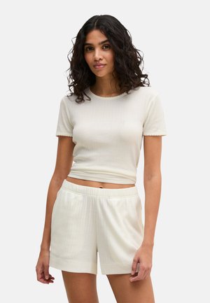 Vrouw met krullend donker haar, gekleed in een crèmekleurige, geribbelde crop top met korte mouwen en bijpassende shorts met elastische taille, staand tegen een effen achtergrond.