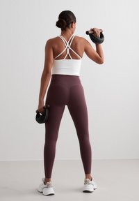 White athletic crop top with crisscross straps, paired with high-waisted maroon leggings and black dumbbells, worn with white sneakers.