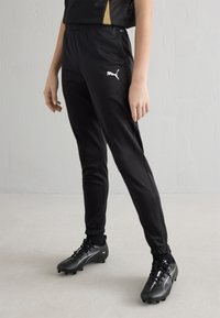 Črne trenirke iz gladkega, lahkega materiala z elastičnim pasom, manšetami in belim logotipom Puma na levi stegni.