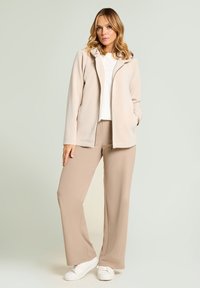 Veste beige clair à fermeture éclair avec capuche et petites décorations, associée à un pantalon beige large assorti et des baskets blanches.